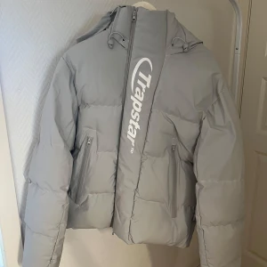 Grå Trapstar pufferjacka med huva - Säljer en grå pufferjacka från Trapstar med stor logga framtill och bak. Jackan har huva med snörning, två fickor med dragkedja och en oversized passform. Materialet är 100% polyester och jackan är perfekt för kalla dagar. Riktigt clean och modern streetstil.