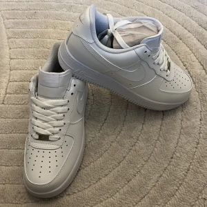 Nike Air Force 1 vita sneakers - Klassiska Nike Air Force 1 sneakers i helvitt skinn med perforerad tå, rund tå och platt sula. Snygg och tidlös modell med snörning och diskret Nike-logga på sidan. Perfekta för dig som vill ha en clean och stilren look.