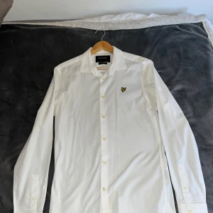 Vit skjorta från Lyle & Scott - Klassisk vit skjorta från Lyle & Scott med gul broderad logga på bröstet. Skjortan har lång ärm, knappar framtill och en stilren krage. Perfekt för dig som gillar en clean och enkel look. Har använts fåtal gånger. Annars enbart legat i garderoben. Mycket bekväm och passar väl till en utekväll eller ett firande. 