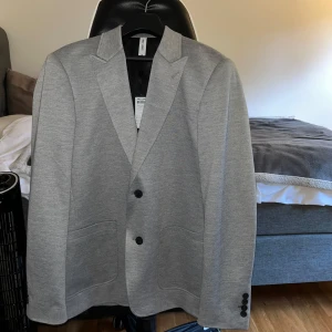 Ljusgrå kavaj från Only & Sons, M - Snygg ljusgrå kavaj från Only & Sons i storlek M. Kavajen har två knappar framtill, klassiska slag och tre knappar vid ärmsluten. Tillverkad i mjuk bomullsblandning med två stora fickor framtill. Perfekt för en stilren look. OBS: Den är helt OANVÄND. Etiketten finns kvar som syns på bilden. Köpt för: 750kr. 