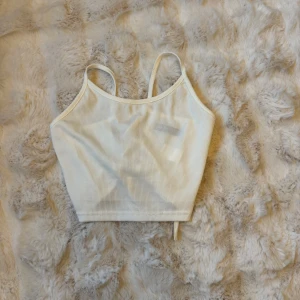 Vit ribbad linnetopp med snörning - Säljer en vit ribbad linnetopp med smala axelband och snygg snörningsdetalj i ryggen. Toppen är croppad och har en stilren, enkel design framtill men cool korsad snörning bak som ger extra edge. Perfekt att styla till jeans eller kjol.