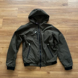 Olivgrön vindjacka från Snoot - Snygg olivgrön vindjacka från Snoot med huva och svarta detaljer. Jackan har tre dragkedjeförsedda fickor framtill och muddar vid ärmslut och midja. Tillverkad i lätt och slitstark polyester, perfekt för blåsiga dagar.