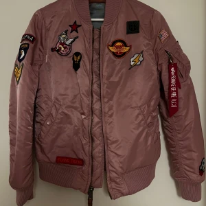 Rosa bomberjacka Alpha Industries M - Säljer en rosa bomberjacka från Alpha Industries med coola patches och detaljer som dragkedja på ärmen och ribbade muddar. Jackan är i glansigt material och har klassisk bomberpassform. 