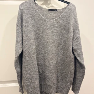 Grå stickad tröja från 157 - Mysig grå stickad tröja från 157 med v-ringad hals och avslappnad passform. Perfekt för lager-på-lager och chill dagar. Långärmad och ribbade muddar vid ärmslut och nederkant. Enkel att matcha med jeans eller kjol.