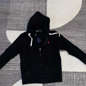 Svart hoodie från Polo Ralph Lauren - Svart hoodie från Polo Ralph Lauren med vit dragsko och dragkedja framtill. Klassisk röd broderad logga på bröstet och känguruficka. Perfekt för chill dagar och snygg streetstyle. Modellen är Slime fit så är du 170-180 så sitter den fint på dig