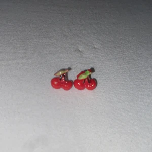 Söta körsbärsörhängen i rött - Små örhängen formade som körsbär med röda bär och gröna blad. Perfekta för dig som vill ha en lekfull och färgglad detalj till din stil. Passar dig som gillar unika accessoarer med fruktmotiv.