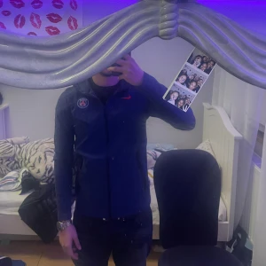 Mörkblå PSG vindjacka från Nike - Säljer en mörkblå vindjacka från Nike med PSG-logga på bröstet och röd Nike-swoosh. Jackan har dragkedja framtill och lång ärm. Perfekt för dig som gillar sportig stil och vill representera Paris Saint-Germain.