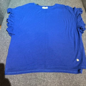 Blå topp med volangärmar från 365 - Snygg blå topp från 365 med korta volangärmar som ger en cool och trendig look. T-shirten har en avslappnad passform och är tillverkad i mjukt material, perfekt för dig som gillar färg och vill sticka ut lite extra.