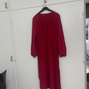 Röd omlottklänning med plisserad kjol - Snygg röd långklänning med omlottliv och djup v-ringning. Klänningen har långa ärmar med lätt puff och plisserad kjol som ger ett flowy intryck. Midjeband för att knyta och markerad midja. Perfekt för dig som vill sticka ut med en elegant look.