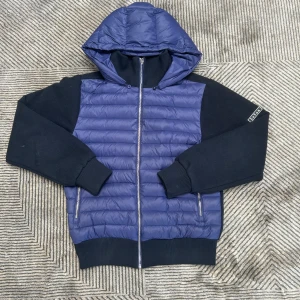 Blå cardigan från LeVon - Säljer en blå och svart pufferjacka från LeVon i storlek XS. Jackan har quiltad framsida, huva och dragkedja hela vägen. Ärmar och rygg är i mjukt tyg med ribbade muddar. Två fickor med dragkedja framtill. Perfekt för kyliga dagar! Den har lite krokig kedja men inget man märker när den är på 