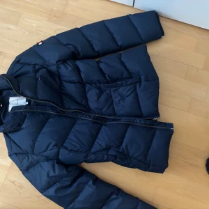 Mörkblå dunjacka Tommy Hilfiger - Snygg mörkblå dunjacka från Tommy Hilfiger med klassisk quiltad design och dragkedja framtill. Jackan har lång ärm, hög krage och diskreta logodetaljer på axeln. Perfekt för kyliga dagar och enkel att matcha med det mesta.