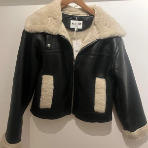 Svart teddyjacka från NA-KD med fuskpäls - Snygg svart teddyjacka från NA-KD med krage, muddar och fickdetaljer i beige fuskpäls. Jackan har dragkedja framtill och är fodrad med mjukt teddyfoder. Perfekt för kalla dagar och ger en cool vibe till din outfit.