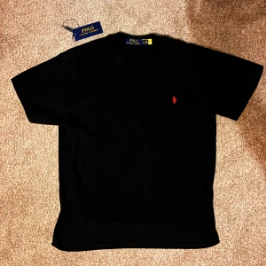 Svart t-shirt från Polo Ralph Lauren - Svart t-shirt från Polo Ralph Lauren med klassisk passform och röd broderad logga på bröstet. Tillverkad i mjuk bomull och har rund halsringning samt korta ärmar. Perfekt för en clean och stilren look.