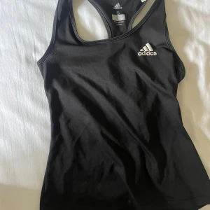 Svart sportlinne från Adidas - Svart sportigt linne från Adidas med klassisk logga på bröstet. Modellen har brottarrygg och är tillverkat i ett lätt och stretchigt material som andas, perfekt för träning. Snygg och enkel design som funkar till alla sportiga outfits.