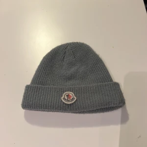 Grå stickad Moncler mössa - Snygg grå ribbstickad mössa från Moncler med klassisk uppvikt kant och broderad logotyp framtill. Mössan har en enkel, rundad form och är tillverkad i mjukt stickat material som håller dig varm. Perfekt för dig som gillar stilrena accessoarer med exklusiv känsla. Den har två små revor som inte syns så mycket men inget som påverkar funktionen.
