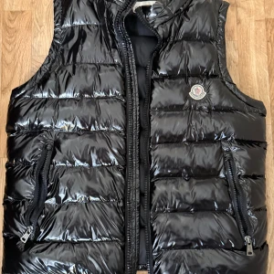 Svart dunväst från Moncler - Snygg svart dunväst från Moncler med glansig finish och klassisk logga på bröstet. Västen har två dragkedjefickor framtill och quiltad design. Perfekt för lager-på-lager och ger en clean streetstyle-vibe. NFC Scan funkar på märket 