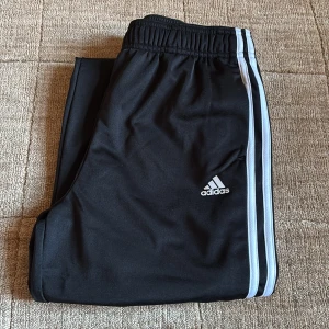 Svarta Adidas träningsbyxor Regular Fit - Svarta träningsbyxor från Adidas med klassiska vita ränder längs sidorna och Adidas-logga på låret. Byxorna har resår i midjan och vid bensluten, samt är tillverkade i ett mjukt och lätt polyestermaterial. Regular fit för en bekväm passform.