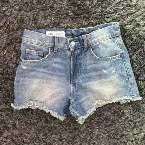 Blå jeansshorts med fransig kant från Zara - Snygga blå jeansshorts från Zara med fransig rå kant och slitna detaljer. Klassisk femficksmodell med bälteshällor och knappstängning. 