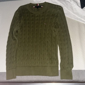 Olivgrön kabelstickad tröja Tommy Hilfiger - Snygg olivgrön kabelstickad tröja från Tommy Hilfiger med rund halsringning och diskret logga på bröstet. Tröjan har ribbade muddar och är långärmad, perfekt för lager-på-lager under hösten och vintern. PRIS KAN DISKUTERAS 