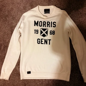 Vit stickad tröja Morris Gents M - Snygg vit stickad tröja från Morris Gents i storlek medium. Tröjan har svart tryck med texten 'Morris Gent 1968' och ett emblem på bröstet. Klassisk rund halsringning och ribbade muddar vid ärmslut och nederkant. Perfekt för en stilren look. Hör av er vid minsta fundering