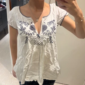 Vit blus med broderi från Odd Molly - Supersöt vit blus från Odd Molly med mörkblå broderade blommor och dekorativt mönster framtill. Blusen har korta ärmar, v-ringning med liten rosett och virkade kanter. Tillverkad i mjuk bomull och har en luftig, avslappnad passform.