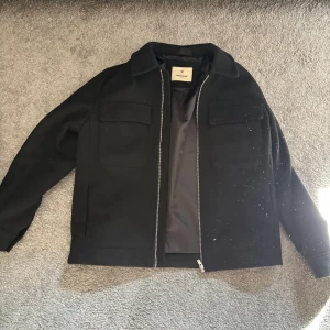 Svart overshirt från Jack&Jones - Svart overshirt från Arket med klassisk krage, två stora bröstfickor och dragkedja framtill. Jackan har en stilren och enkel design som passar till många olika outfits. Materialet är ullblandning som ger en mjuk och varm känsla.