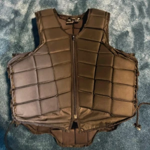 Svart skyddsväst från Horse Life - Skyddsväst från Horse Life i svart med quiltad design och dragkedja framtill. Västen har vadderade paneler för extra skydd och snörning i sidorna för justerbar passform. Perfekt för ridning och andra aktiviteter där extra skydd behövs. Aldrig varit med om något fall