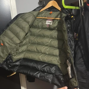 Svart dunväst från Parajumpers  - Säljer en svart dunväst från Parajumpers, modell Sheen. Västen har quiltad design, dragkedja framtill och coola detaljer som märkets patch på insidan. Insidan är olivgrön och materialet är glansigt och slitstarkt. Perfekt för lager-på-lager under höst och vinter jacka