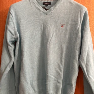 Ljusblå stickad tröja från GANT - Snygg ljusblå stickad tröja från GANT med v-ringning och broderad logga på bröstet. Tröjan har ribbade muddar och är långärmad, perfekt för lager-på-lager under kyliga dagar.