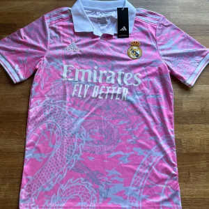 Real Madrid Fotbollströja  - Snygg Real Madrid fotbollströja från Adidas i rosa med ljusgrått drakmönster. Tröjan har vit krage, korta ärmar och klassiska Adidas-ränder på axlarna. Materialet är lätt och andas, perfekt för match eller träning. Officiell klubbmärke och sponsortryck på bröstet.