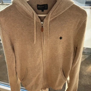 Morris tröja - Säljer en beige hoodie från Morris i stickad bomull med dragkedja och huva. Tröjan har snörning vid huvan, två fickor framtill och en liten broderad logga på bröstet. Perfekt för dig som gillar en clean och avslappnad stil. Använd väldigt varsamt. Pris går att diskuteras!