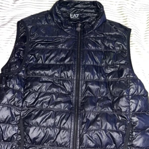 Svart dunväst från EA7 Emporio Armani - Snygg svart dunväst från EA7 Emporio Armani med quiltad design och hög krage. Västen har en tydlig logga på bröstet och dragkedja med EA7-märke. Perfekt att slänga på över en hoodie för en cool look. Materialet är glansigt och lätt, vilket gör den enkel att bära. Du kan göra sjuka outfits med den. (Ny pris 1 751kr) 