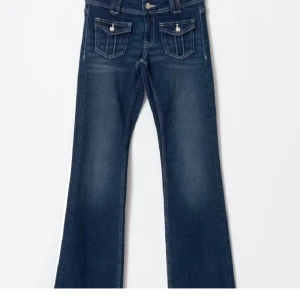Blå bootcut jeans med fickdetaljer - Snygga blå jeans med bootcut passform och markerade fickor framtill med lock och knapp. Jeansen har klassisk femficksdesign och coola slitningar för en trendig look. Perfekta för dig som gillar en avslappnad men ändå stilren vibe.