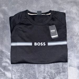 Svart långärmad tröja från BOSS - Svart långärmad tröja från BOSS med stilren logga och två gråa ränder över bröstet. Tröjan har rund halsringning och ribbade muddar vid ärmslut och nederkant. Tillverkad i mjuk bomull för en skön känsla. Tröjan är köpt i somras och har aldrig använts. OBS: Etikett är kvar. Den kom lite defekt med ett litet snitt längst ned på ärmen som bilden visar. Annars helt ny. Originalpris: 700kr. 
