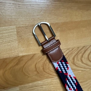 Randigt flätat bälte med skinn - Säljer ett snyggt flätat bälte med randigt mönster i marinblått, vitt och rött. Spännet är i silverfärgad metall och ändarna är förstärkta med brunt skinn. Perfekt för att ge outfiten lite extra edge.