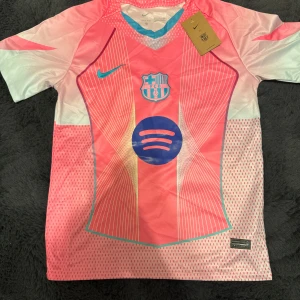 FC Barcelona x Nike rosa fotbollströja - Snygg FC Barcelona fotbollströja från Nike i rosa och ljusblått med vita detaljer. Tröjan har korta ärmar, Dri-FIT-material och ett unikt grafiskt mönster med klubbmärke och Spotify-logga på bröstet. Perfekt för dig som vill sticka ut på planen.