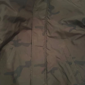 Camouflage vindjacka från H&M Divided XS - Säljer en snygg vindjacka från H&M Divided i storlek XS. Jackan har ett klassiskt camouflage-mönster i mörka gröna och bruna toner, huva och två snedställda fickor framtill. Perfekt för dig som vill ha en streetig look.