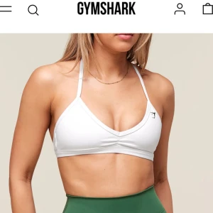 Vit sport topp från Gymshark - Nästan aldrig använd, Vit sport topp fårn  Gymshark med smala axelband som korsas i ryggen. 💕Toppen har en enkel och stilren design med liten svart logga framtill. Perfekt för träning och har en tight passform som sitter snyggt på kroppen. Pris går att diskutera💗