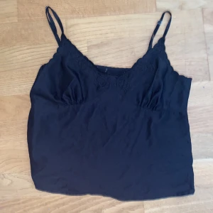 Svart linnetopp med spetsdetaljer - Svart linnetopp med tunna axelband och v-ringning. Toppen har broderade spetsdetaljer med blommönster längs bysten och en lätt croppad passform. Jättefin till någon simpel fest eller tjejkväll 💞