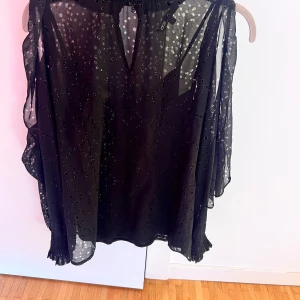 Svart glittrig kortklänning från Monki - Svart blus från Maje i transparent, lätt material med glittriga prickar. Blusen har långa ärmar med volangdetaljer och smock vid ärmslut. Det är även öppet vid axlarna som gör den ännu coolare. Perfekt för dig som gillar en trendig och lekfull stil.