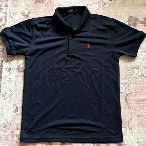 Svart pikétröja från Ralph Lauren - Klassisk svart pikétröja från Ralph Lauren med röd broderad logga på bröstet. Tröjan har krage, korta ärmar och knäppning framtill. Tillverkad i bomull för en skön och luftig känsla. Perfekt för en clean och stilren look.