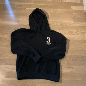 Svart hoodie från Vinson Polo Club - Svart hoodie från Vinson Polo Club med huva och snörning. Tryck med siffran 3 och Polo Club på bröstet. Snygga detaljer med logoband i huvan. Har några mindre skavanker.