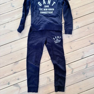 Mörkblå sweatshirt och byxa från GANT - Mörkblå sweatshirt samt byxa från GANT med vit tryckt logga och text på bröstet. Tröjan har rund hals, ribbade muddar och långa ärmar. Perfekt för chill dagar och har en klassisk, sportig vibe. Materialet är mjuk bomull som känns skönt mot huden. Passar både tjej och kille. 134/140