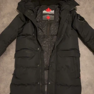 Svart lång dunjacka från Zavetti Canada - Säljer en svart lång dunjacka från Zavetti Canada med huva och flera fickor. Jackan har dragkedja och tryckknappar framtill, samt snygga detaljer som broderat lönnlöv på ärmen och ryggen. Perfekt för kalla dagar och riktigt stilren look.