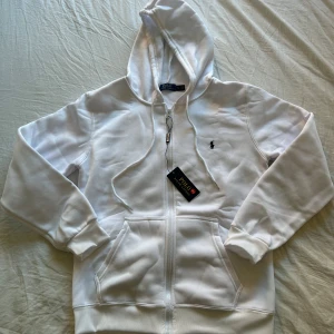 Vit hoodie från Polo Ralph Lauren - Vit hoodie från Polo Ralph Lauren med dragkedja, snörning i huvan och klassisk broderad logga på bröstet. Tröjan har två fickor framtill och ribbade muddar vid ärmslut och nederkant. Perfekt för chill dagar och enkel att matcha.