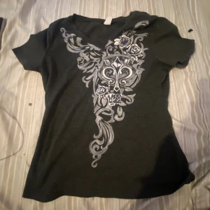 Svart t-shirt med silvrigt tryck - Svart t-shirt med korta ärmar och snyggt silvrigt tryck framtill med blommor och grafiska detaljer. T-shirten har en klassisk passform och är gjord i mjukt material, perfekt för en cool och avslappnad stil.