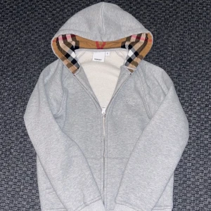 Grå hoodie från Burberry med rutig huva - Snygg grå hoodie från Burberry med dragkedja och klassiskt broderat logomärke på bröstet. Huvan är fodrad med ikoniskt rutigt Burberry-tyg i beige, rött och svart. Perfekt för dig som vill ha en stilren och exklusiv look.pris kan diskuteras!