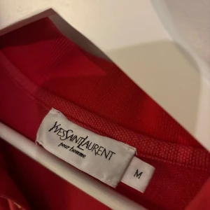 Röd pikétröja från Yves Saint Laurent - Röd pikétröja från Yves Saint Laurent med klassisk krage, knäppning med tre knappar och broderad YSL-logga på bröstet. Tröjan har korta ärmar och är tillverkad i mjuk bomull som känns skön mot huden. Piken har en sitter lite löst på kroppen och kan passa både L och XL. Den har en fin kvalité och vill därför inte pruta med priset.