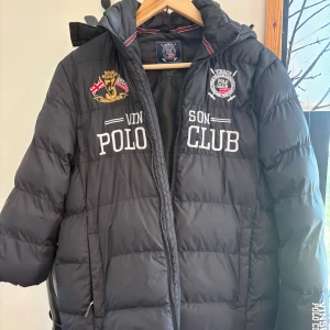 Svart pufferjacka från Vinson Polo Club 160 storlek  - Svart pufferjacka från Vinson Polo Club med broderade loggor och text på bröstet. Jackan har huva, dragkedja framtill och quiltad design. Perfekt för kalla dagar och har en sportig vibe med snygga detaljer.