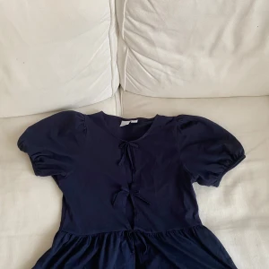 Mörkblå topp/blus från Gina tricot - Supersöt mörkblå topp från Gina Tricot med puffärmar och tre knytningar framtill. Modellen är kort och har en lätt utställd kjol. Tillverkad i mjuk bomull som känns skön mot huden. Perfekt för dig som gillar en enkel men ändå trendig look. Änvänd en gång i storlek 158 164
